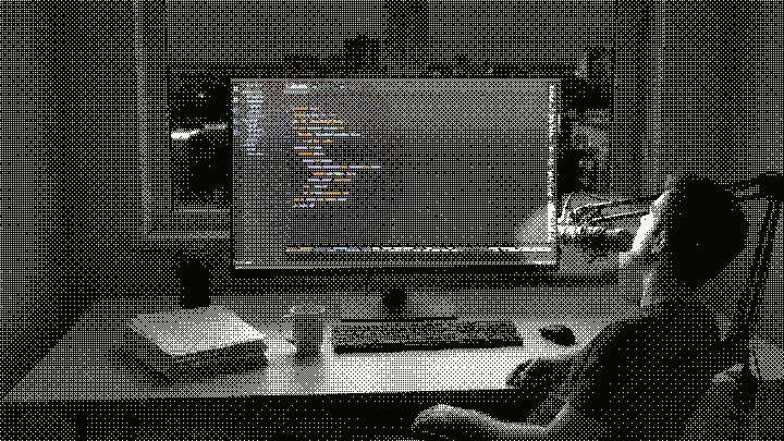 The same desk, the same room, the same mug and plant, at night, from the same place over the shoulder. The dots have inverted to light on black. The person has sat back with both hands away from the keyboard and mouse, head up, speaking into a microphone on a boom arm. The same editor is on the monitor, now down to a single tidy column of code in colour with empty space around it.